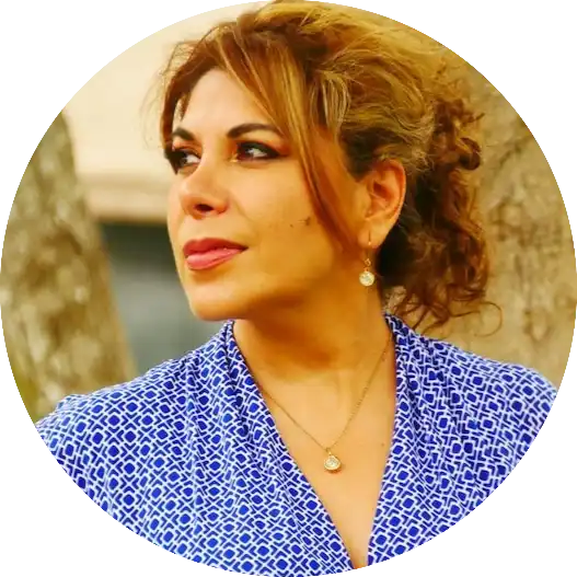 A woman with auburn hair, wearing a blue and white patterned shirt, a gold necklace, and gold earrings.
