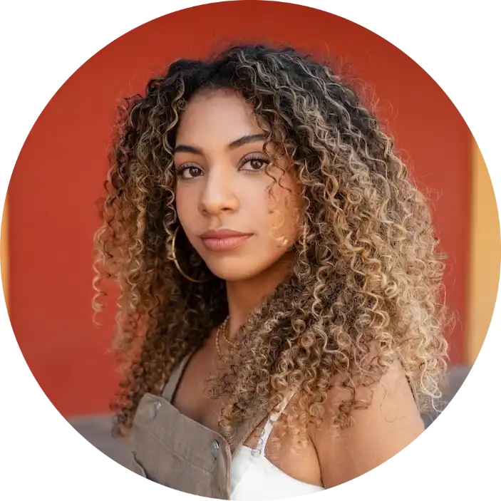 A young woman with long, curly, highlighted hair and hoop earrings, wearing a white top and overalls.